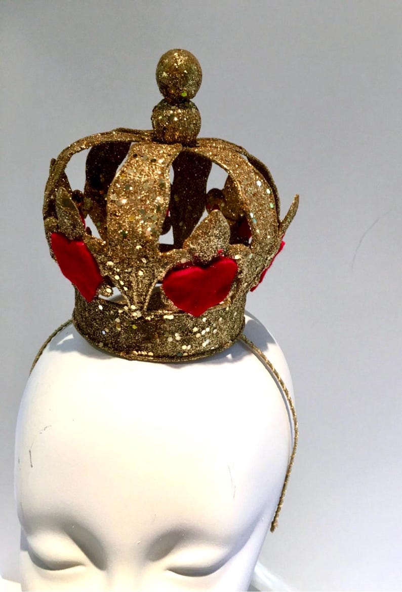 May include: A gold glitter crown headband featuring a series of raised red heart-shaped accents around the base. The crown has a central spherical finial at the top and is attached to a thin, matching gold glitter-covered headband frame.