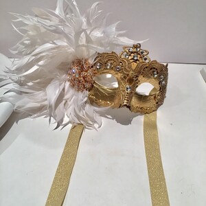 May include: A gold masquerade mask with white feathers and rhinestones. The mask has a delicate lace design and is attached to two gold ribbons.