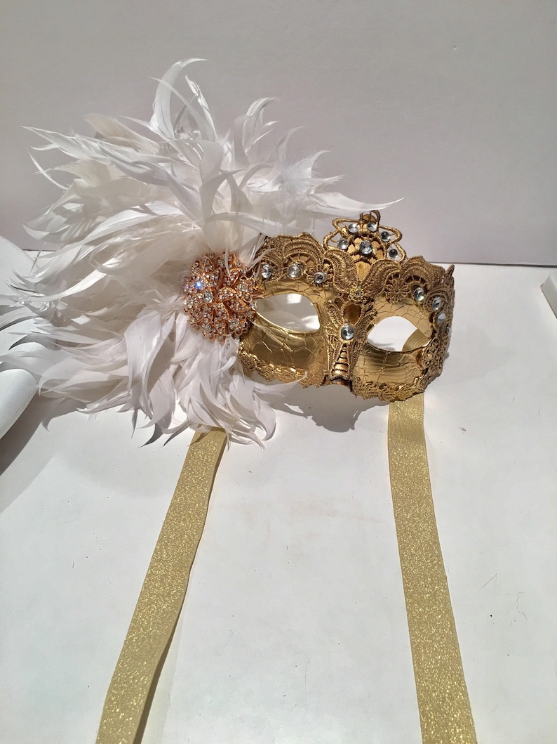 May include: A gold masquerade mask with white feathers and rhinestones. The mask has a delicate lace design and is attached to two gold ribbons.