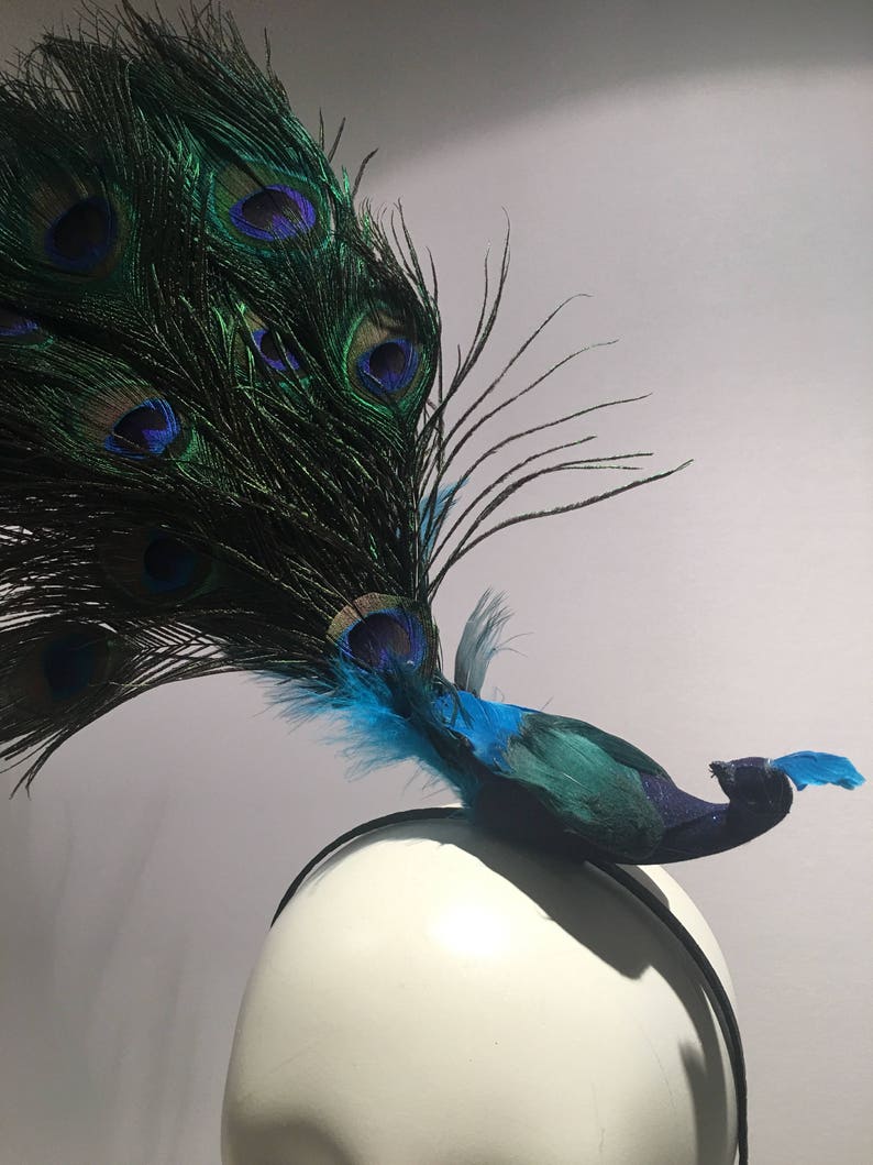 May include: A peacock feather headband with a blue and green peacock feather design. The headband is black and is on a white mannequin head.