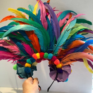 May include: A colorful feather headband with rainbow pom poms and purple flowers. The headband is made of black elastic and is decorated with feathers in various colors, including blue, green, yellow, orange, pink, and red. The headband is perfect for a festival or party.