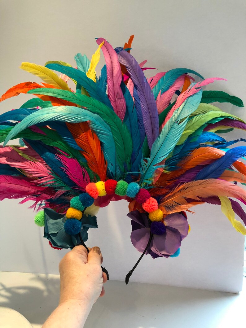 May include: A colorful feather headband with rainbow pom poms and purple flowers. The headband is made of black elastic and is decorated with feathers in various colors, including blue, green, yellow, orange, pink, and red. The headband is perfect for a festival or party.
