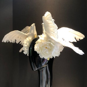 May include: A decorative headpiece featuring white doves with outstretched wings, perched atop a black mannequin head. The headpiece is adorned with white flowers and feathers, creating a dramatic and elegant design.