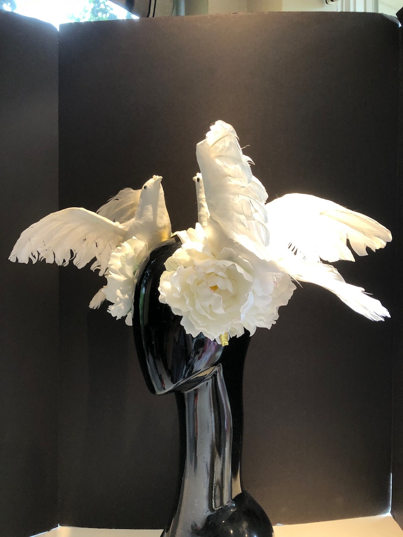 May include: A decorative headpiece featuring white doves with outstretched wings, perched atop a black mannequin head. The headpiece is adorned with white flowers and feathers, creating a dramatic and elegant design.