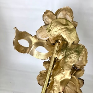 K&ouml;nnte beinhalten: Eine goldfarbene Maske f&uuml;r einen Maskenball mit einem Blumenarrangement. Die Maske ist mit kleinen, dekorativen Perlen an den R&auml;ndern verziert. Die Blumen und Bl&auml;tter sind ebenfalls goldfarben und bilden ein stimmiges, elegantes Design.