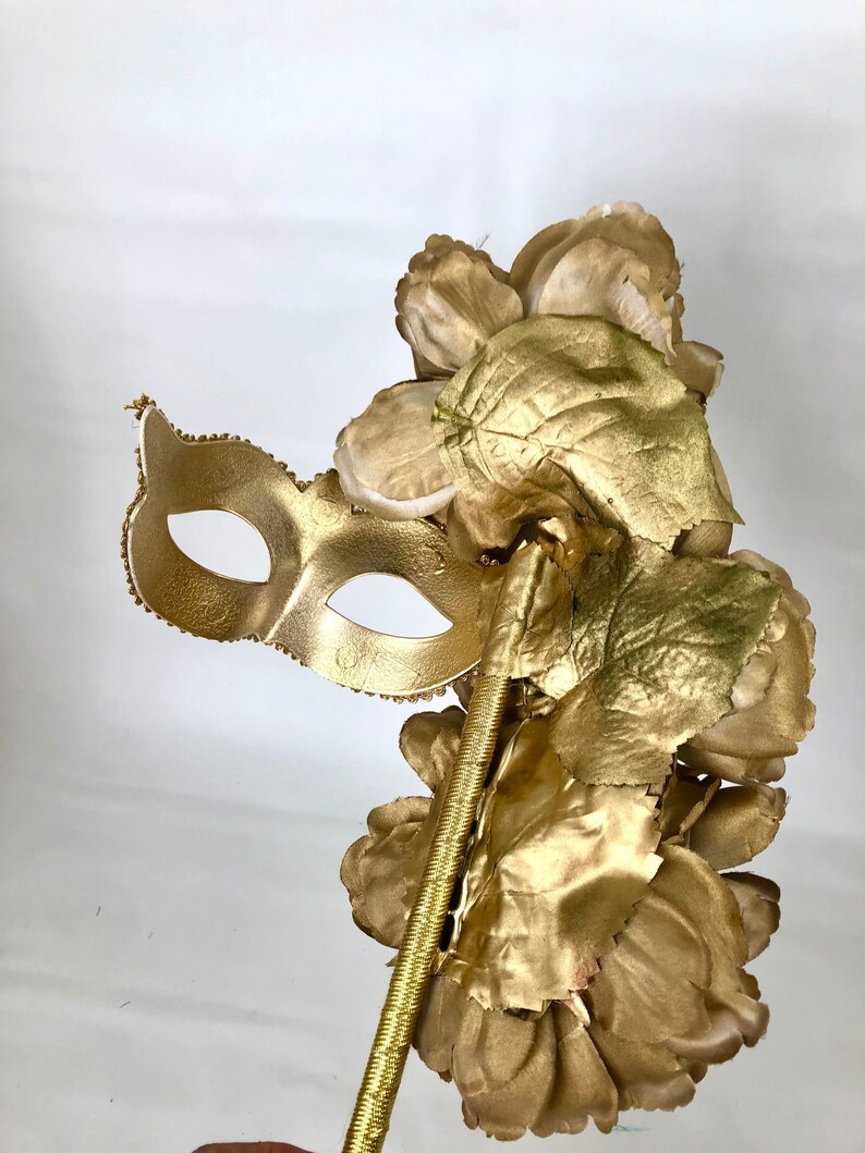 K&ouml;nnte beinhalten: Eine goldfarbene Maske f&uuml;r einen Maskenball mit einem Blumenarrangement. Die Maske ist mit kleinen, dekorativen Perlen an den R&auml;ndern verziert. Die Blumen und Bl&auml;tter sind ebenfalls goldfarben und bilden ein stimmiges, elegantes Design.