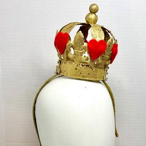 May include: A gold glitter crown headband featuring a series of red felt heart cutouts attached to the exterior points. The crown has a central spherical finial at the top and is mounted on a thin, matching gold glitter-covered headband frame.