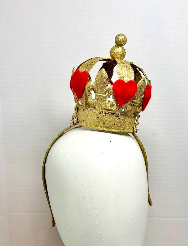 May include: A gold glitter crown headband featuring a series of red felt heart cutouts attached to the exterior points. The crown has a central spherical finial at the top and is mounted on a thin, matching gold glitter-covered headband frame.