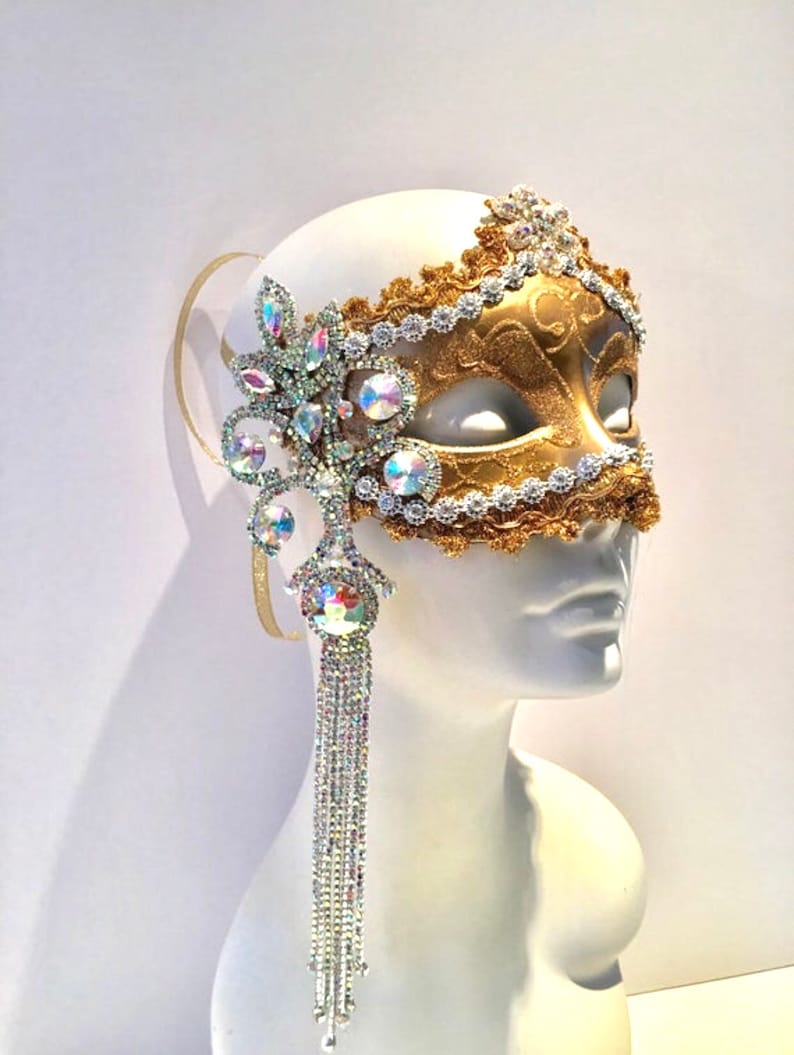 May include: A gold masquerade mask with a white lace trim and a crystal embellishment. The mask has a long crystal fringe hanging from the side.