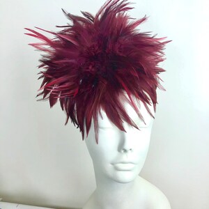 K&ouml;nnte beinhalten: Ein Kopfschmuck aus burgunderfarbenen Federn mit einem gro&szlig;en, flauschigen Design. Der Kopfschmuck befindet sich auf einem wei&szlig;en Mannequin-Kopf.