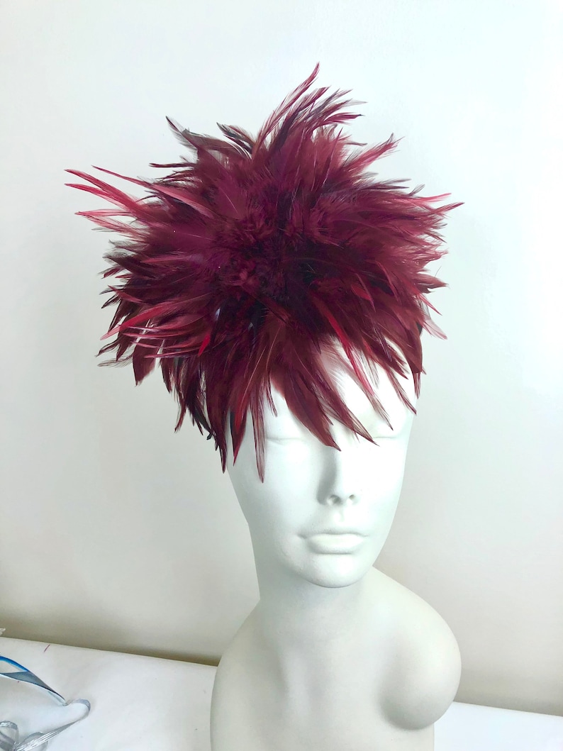 K&ouml;nnte beinhalten: Ein Kopfschmuck aus burgunderfarbenen Federn mit einem gro&szlig;en, flauschigen Design. Der Kopfschmuck befindet sich auf einem wei&szlig;en Mannequin-Kopf.