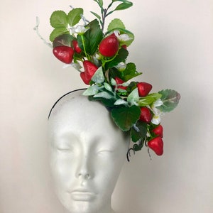 以下が含まれることがあります： イチゴと花の装飾が施された黒いヘアバンド。明るい赤色のイチゴ、緑の葉、小さな白い花が特徴です。ヘッドピースは白いマネキンの頭に飾られています。