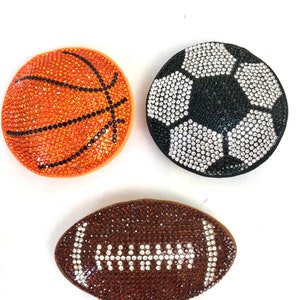 May include: Three sports-themed decorative items: a basketball, a soccer ball, and a football. The basketball is orange with black lines, the soccer ball is black and white, and the football is brown with white details. All are covered in sparkling rhinestones.