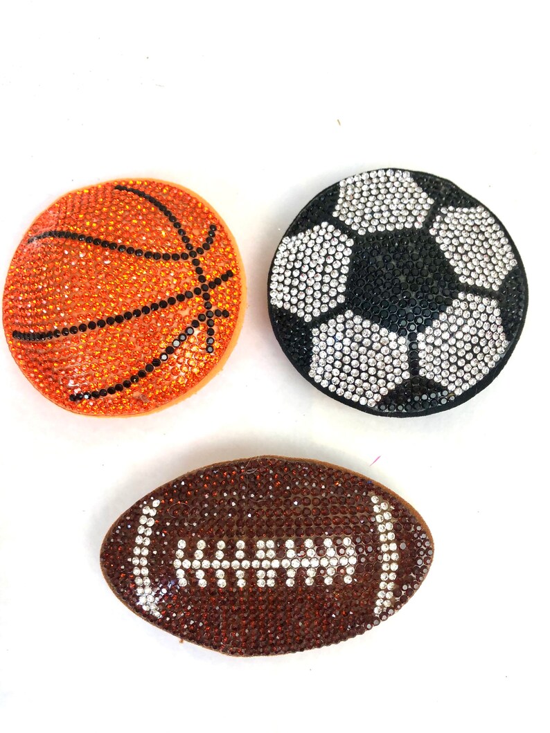 May include: Three sports-themed decorative items: a basketball, a soccer ball, and a football. The basketball is orange with black lines, the soccer ball is black and white, and the football is brown with white details. All are covered in sparkling rhinestones.