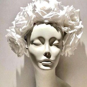 May include: A white floral headband featuring a dense arrangement of large, layered white rose blossoms. The accessory is displayed on a smooth, featureless white mannequin head, highlighting the uniform white color and the intricate petal structure.