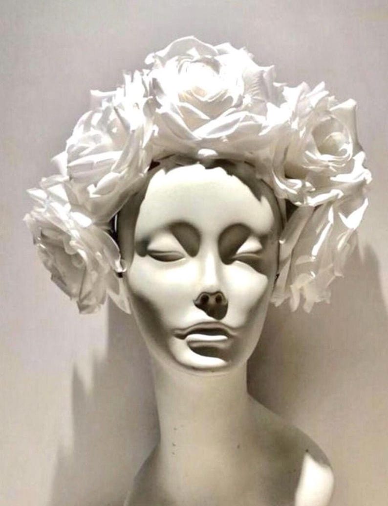 May include: A white floral headband featuring a dense arrangement of large, layered white rose blossoms. The accessory is displayed on a smooth, featureless white mannequin head, highlighting the uniform white color and the intricate petal structure.
