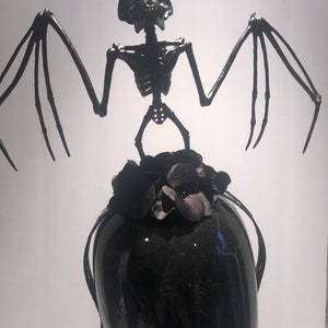 May include: A black decorative sculpture featuring a bat skeleton with outstretched wings atop a black vase. The bat has detailed skeletal features and the vase is adorned with black floral accents. The sculpture is set against a white background.