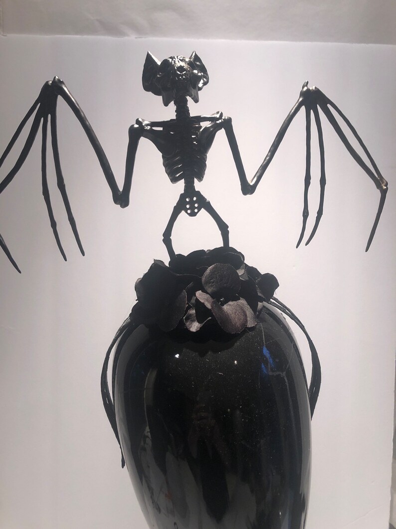 May include: A black decorative sculpture featuring a bat skeleton with outstretched wings atop a black vase. The bat has detailed skeletal features and the vase is adorned with black floral accents. The sculpture is set against a white background.