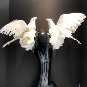 May include: A decorative headpiece featuring two white dove figures with outstretched wings. The doves are adorned with white feathers and are positioned on a black base, accented with white flowers. The headpiece is designed for special occasions.