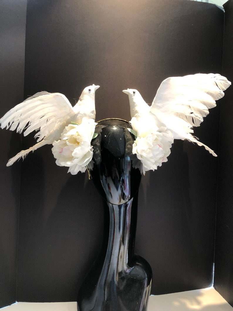 May include: A decorative headpiece featuring two white dove figures with outstretched wings. The doves are adorned with white feathers and are positioned on a black base, accented with white flowers. The headpiece is designed for special occasions.