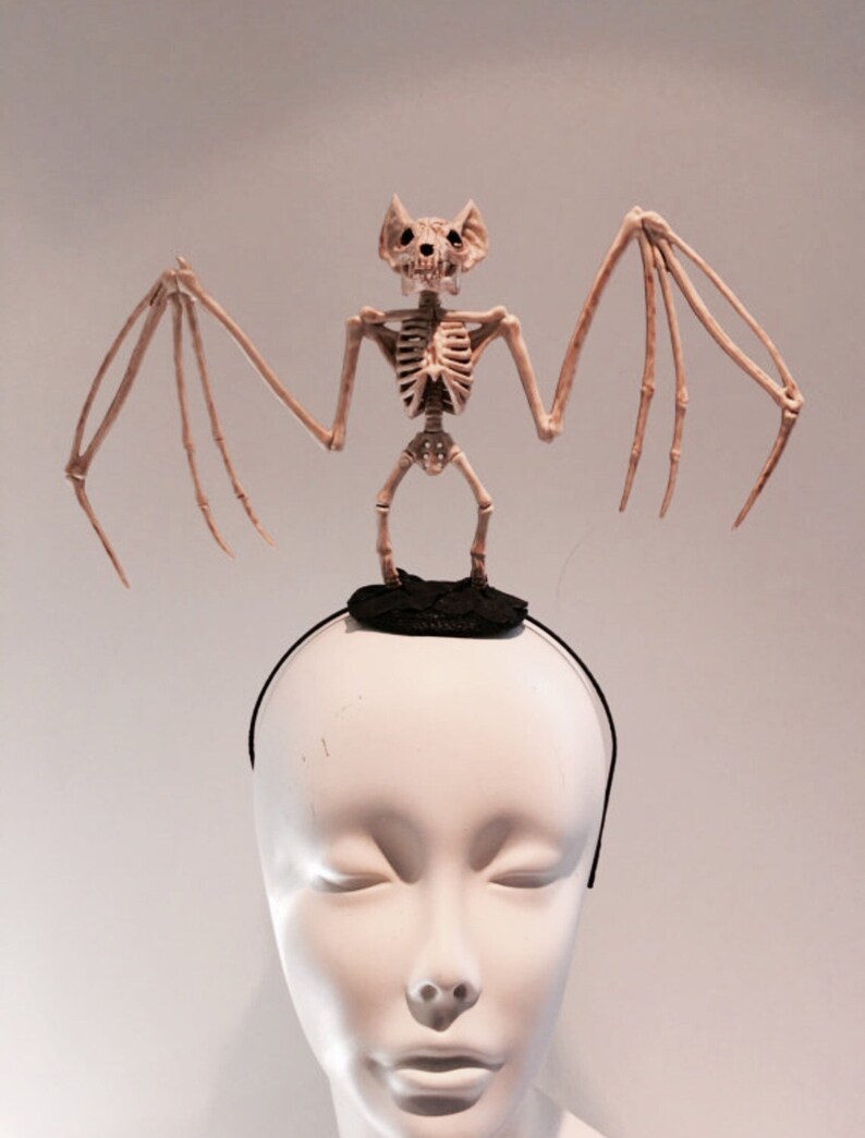 May include: A Halloween-themed headband featuring a skeletal bat figure. The bat is beige and stands atop a small black platform attached to a black headband. The figure has outstretched wings and a detailed skull.