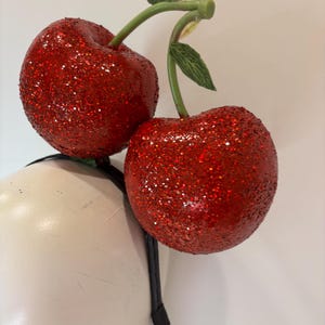 K&ouml;nnte beinhalten: Ein festliches Stirnband mit zwei gro&szlig;en, glitzerbedeckten roten Kirschen. Die Kirschen sind an gr&uuml;nen Stielen mit Bl&auml;ttern befestigt. Das Stirnband ist schwarz.