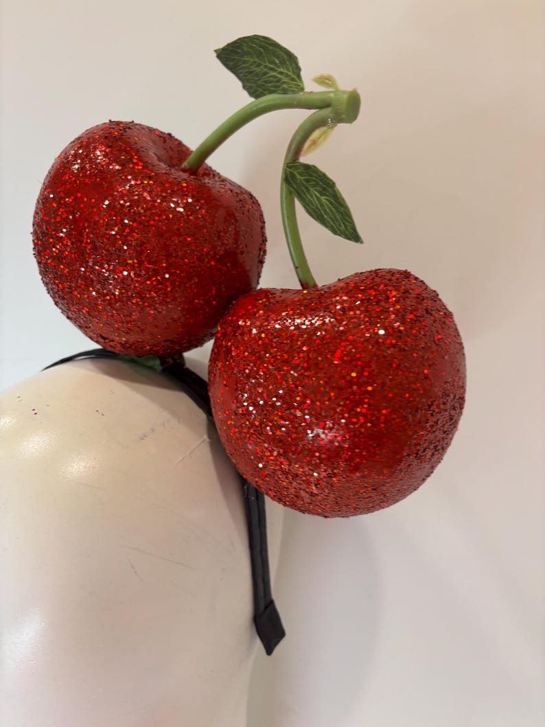 K&ouml;nnte beinhalten: Ein festliches Stirnband mit zwei gro&szlig;en, glitzerbedeckten roten Kirschen. Die Kirschen sind an gr&uuml;nen Stielen mit Bl&auml;ttern befestigt. Das Stirnband ist schwarz.