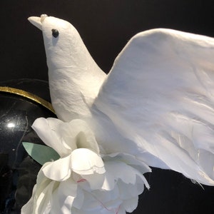 May include: A white dove figurine with outstretched wings, adorned with a white peony flower. The dove has a pearl on its beak and is set against a black background. The dove's wings are made of feathers.