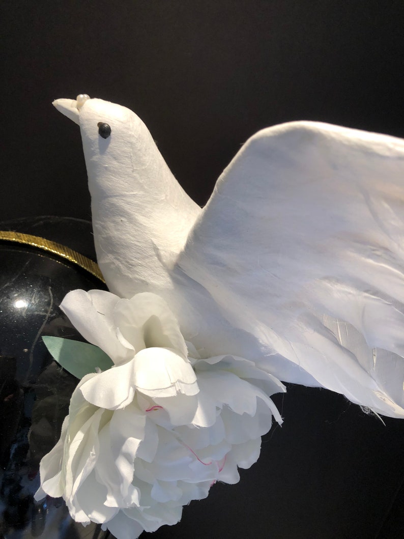 May include: A white dove figurine with outstretched wings, adorned with a white peony flower. The dove has a pearl on its beak and is set against a black background. The dove's wings are made of feathers.