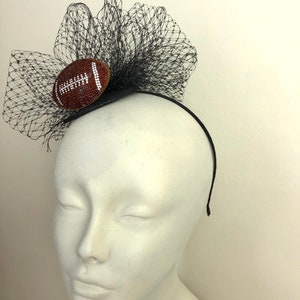 May include: A black headband with a decorative football and black netting. The football is brown with white stitching and embellished with small, sparkling crystals. The netting is layered and adds volume to the design.