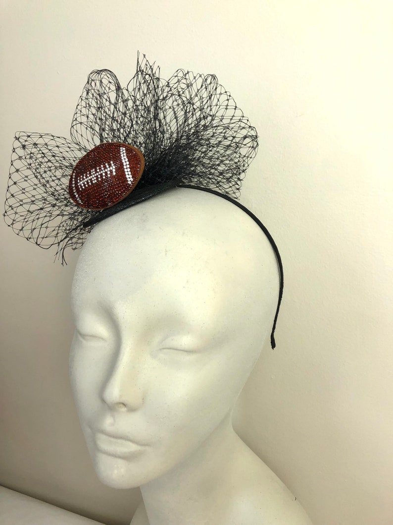 May include: A black headband with a decorative football and black netting. The football is brown with white stitching and embellished with small, sparkling crystals. The netting is layered and adds volume to the design.