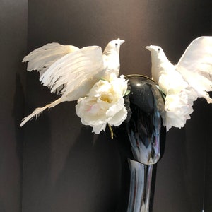 May include: A decorative headpiece featuring two white dove figurines with outstretched wings, perched atop a black mannequin head. The doves are surrounded by white peony flowers and a gold-colored band.