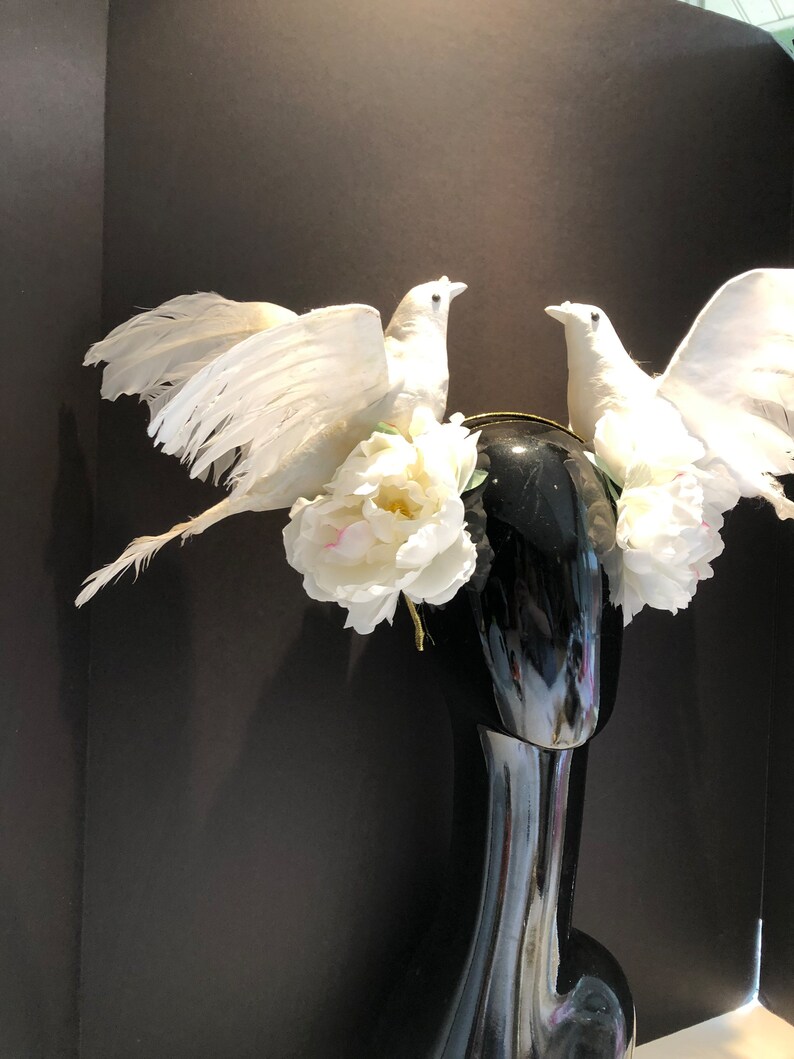 May include: A decorative headpiece featuring two white dove figurines with outstretched wings, perched atop a black mannequin head. The doves are surrounded by white peony flowers and a gold-colored band.