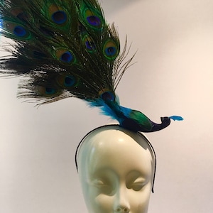 May include: A peacock feather headband with a blue and green peacock figurine. The headband is black and the peacock has a blue body with green feathers.