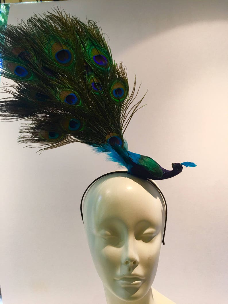 May include: A peacock feather headband with a blue and green peacock figurine. The headband is black and the peacock has a blue body with green feathers.