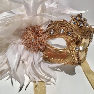 May include: A gold masquerade mask with white feathers and rhinestones. The mask has a delicate lace design and is attached to a gold ribbon.