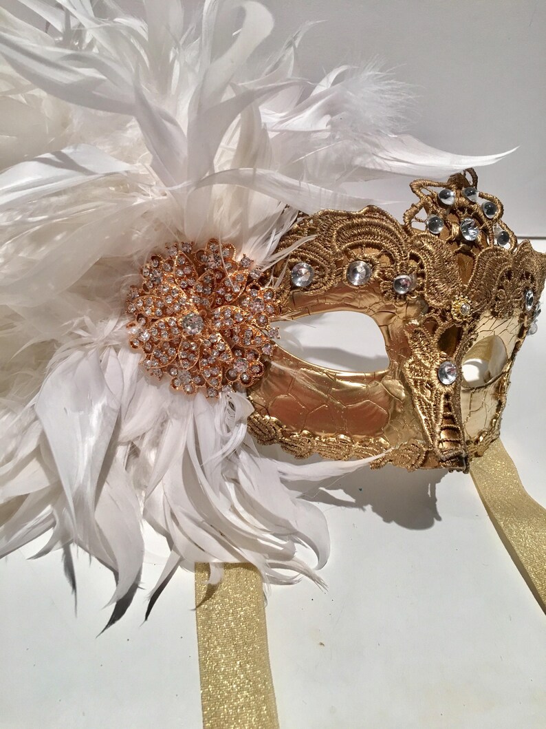 May include: A gold masquerade mask with white feathers and rhinestones. The mask has a delicate lace design and is attached to a gold ribbon.
