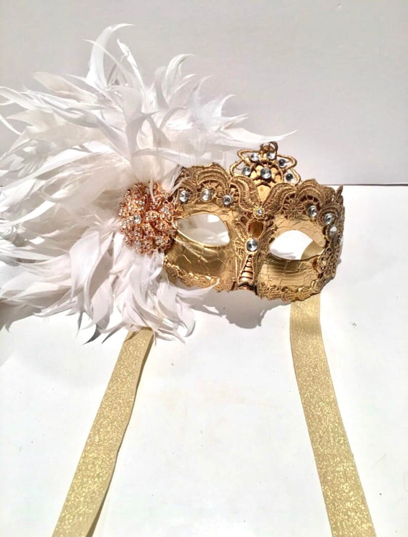May include: A gold masquerade mask with white feathers and a gold ribbon. The mask has intricate details and rhinestones.