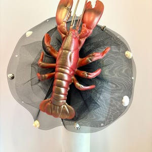 Op de afbeelding: Een zwarte tule hoed met een rode en gouden kreeft erop. De hoed is versierd met kleine witte schelpen.