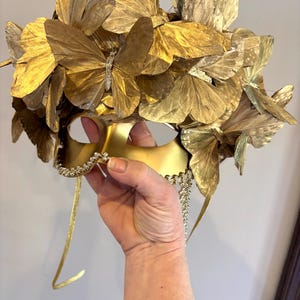 Puede incluir: Una m&aacute;scara dorada para un baile de m&aacute;scaras con una corona de mariposas. La m&aacute;scara est&aacute; adornada con numerosas mariposas doradas y un ribete decorativo de pedrer&iacute;a. La m&aacute;scara tiene lazos dorados.
