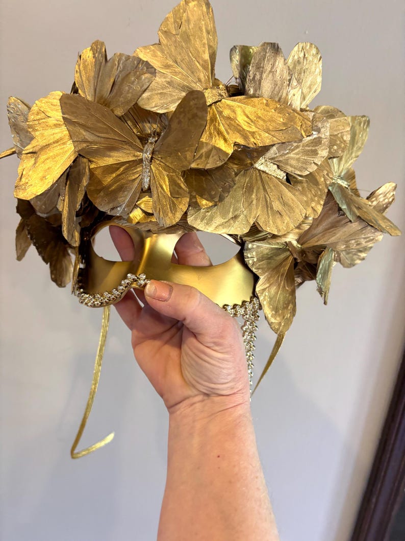Puede incluir: Una m&aacute;scara dorada para un baile de m&aacute;scaras con una corona de mariposas. La m&aacute;scara est&aacute; adornada con numerosas mariposas doradas y un ribete decorativo de pedrer&iacute;a. La m&aacute;scara tiene lazos dorados.