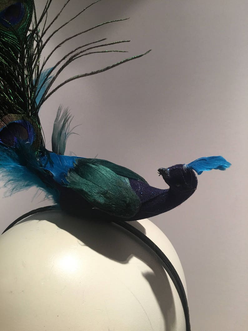 May include: A peacock headband with blue and green feathers. The peacock is perched on a black headband.