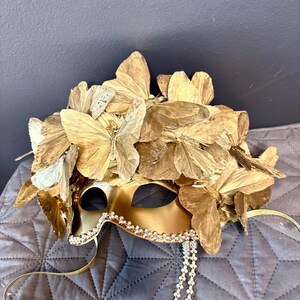 Puede incluir: M&aacute;scara dorada para mascarada con detalles de mariposas. La m&aacute;scara est&aacute; adornada con numerosas alas de mariposa doradas y un ribete de peque&ntilde;os cristales brillantes. Una delicada cadena de cristales cuelga de la m&aacute;scara.
