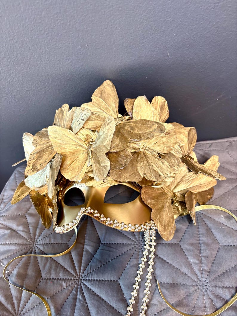 Puede incluir: M&aacute;scara dorada para mascarada con detalles de mariposas. La m&aacute;scara est&aacute; adornada con numerosas alas de mariposa doradas y un ribete de peque&ntilde;os cristales brillantes. Una delicada cadena de cristales cuelga de la m&aacute;scara.