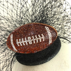 May include: A black fascinator with a football-shaped embellishment. The football is brown and covered in orange and white rhinestones. A black netting and feathers complete the design.