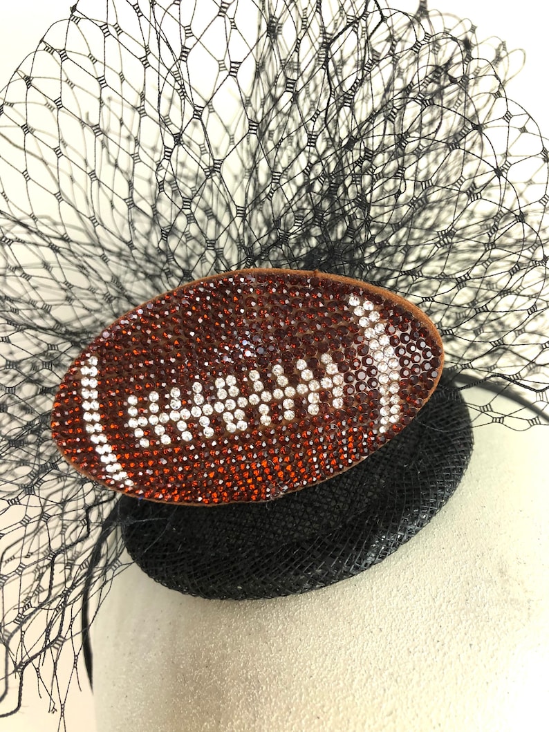 May include: A black fascinator with a football-shaped embellishment. The football is brown and covered in orange and white rhinestones. A black netting and feathers complete the design.