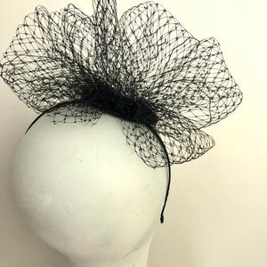 May include: A black headpiece featuring a net-like design, mounted on a thin black headband. The net is shaped into a large, decorative flower, suitable for formal events or costume use.