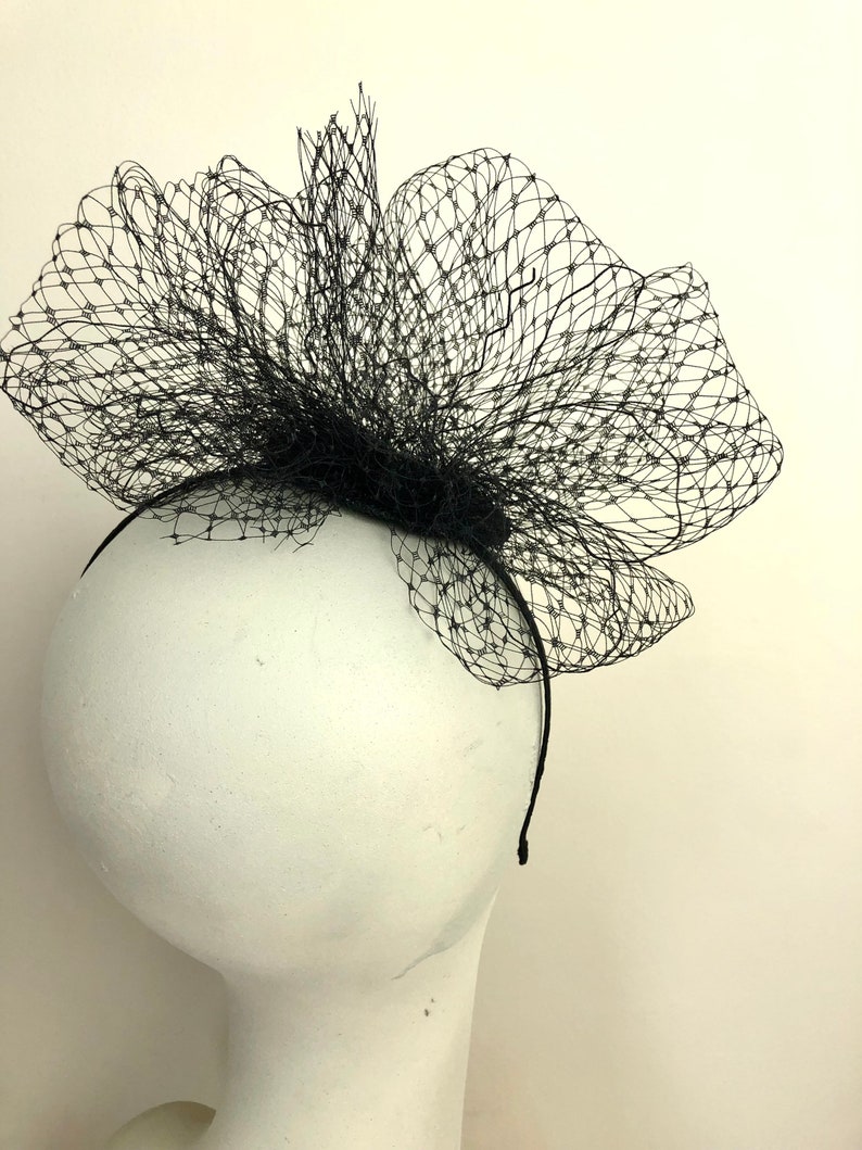 May include: A black headpiece featuring a net-like design, mounted on a thin black headband. The net is shaped into a large, decorative flower, suitable for formal events or costume use.