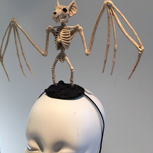May include: A Halloween-themed headband featuring a skeletal bat figure. The bat is beige and stands atop a small black circular base. The headband itself is a thin black band, designed to be worn on the head. The bat's wings are outstretched.