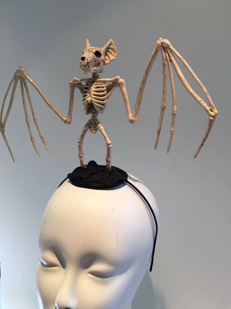 May include: A Halloween-themed headband featuring a skeletal bat figure. The bat is beige and stands atop a small black circular base. The headband itself is a thin black band, designed to be worn on the head. The bat's wings are outstretched.